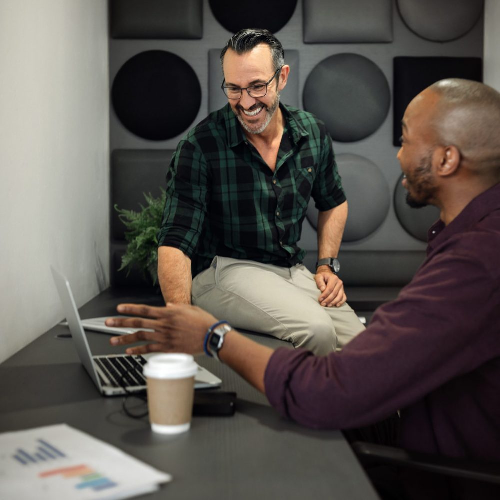 Two smiling diverse businessmen talking together over a laptop while sitting at a desk inside of an office cubicle