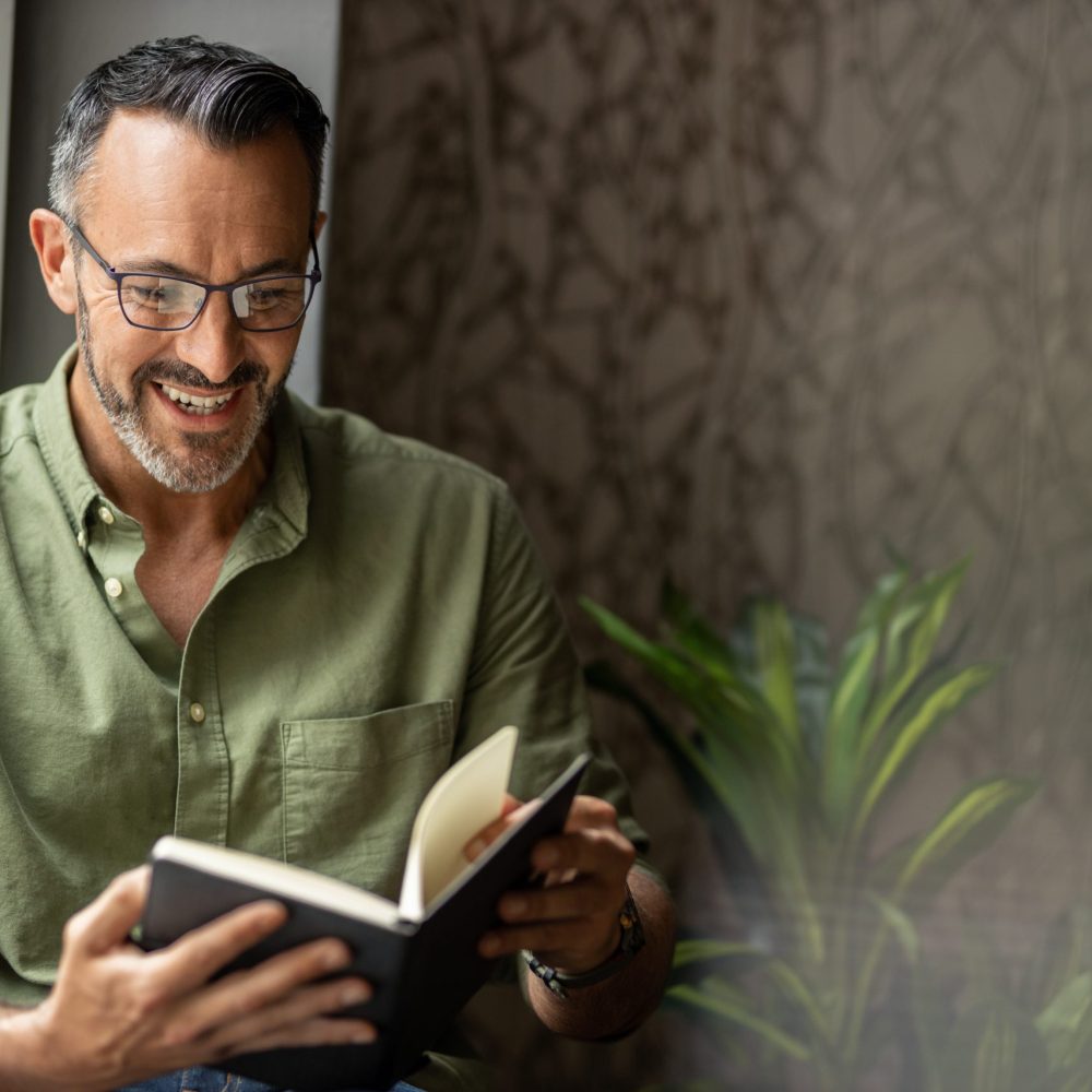 Mature bearded man holding and reading book. Sitting next to sunny window, smiling and wearing glasses, copy space